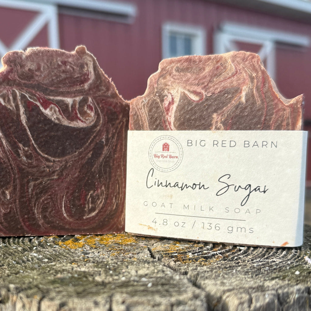 Two bars of cinnamon-scented goat milk soap with a label in front of a red barn.