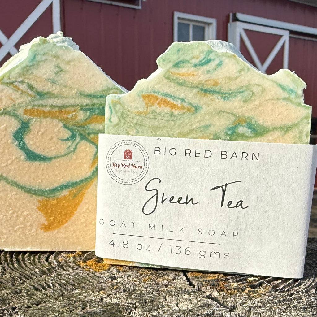 Two bars of green tea goat milk soap with a label in front of a red barn.